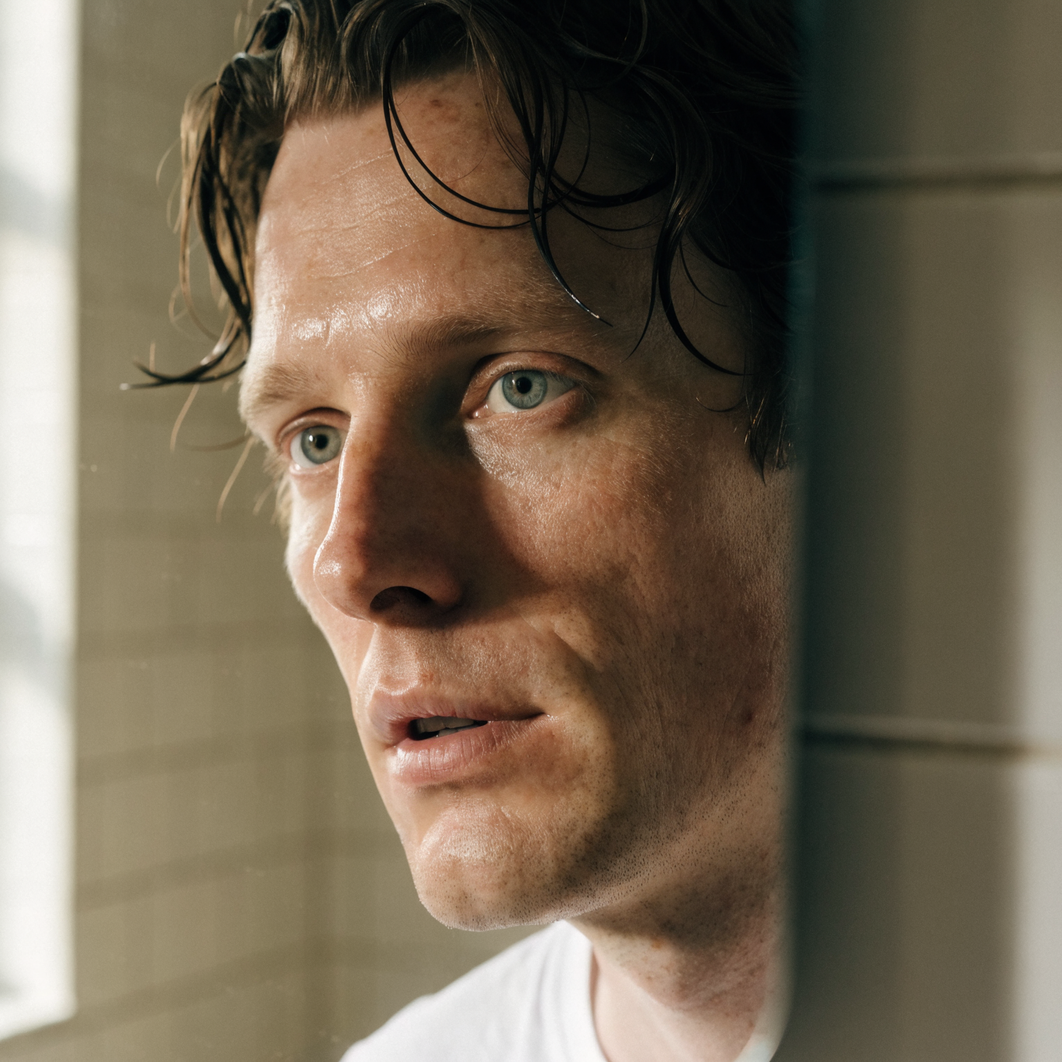 A man looking at his bare, freshly washed face in a bathroom mirror in morning light
